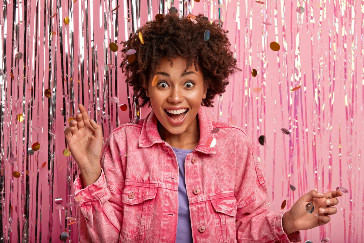 Indoor shot of happy surprised dark skinned lady with Afro haircut, dressed in pink fashionable jacket, poses over confetti and tinsel background, being on bash, feels surprised to be invited on dance